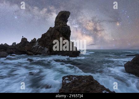 Paesaggio suggestivo che mostra la via Lattea sparsa su tumultuose onde del mare che si infrangono contro una maestosa formazione rocciosa, con una persona solitaria st Foto Stock