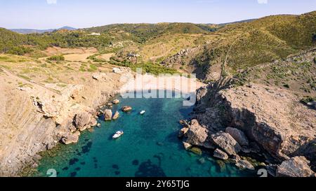 Splendida ripresa aerea di Cala Caldarer, Minorca, che mostra le sue acque turchesi, una spiaggia di sabbia isolata circondata da scogliere rocciose e barche galleggianti Foto Stock
