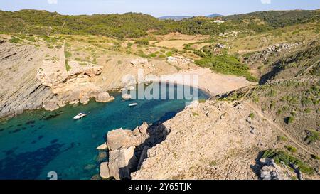 Questa immagine aerea cattura la tranquilla bellezza di Cala Caldarer a Minorca, con acque azzurre trasparenti, barche ancorate e l'aspra costa Foto Stock