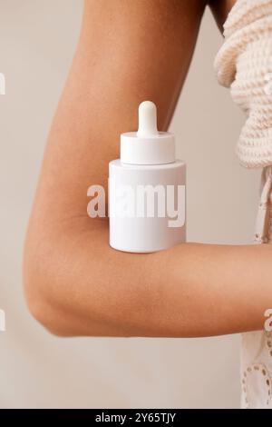 Immagine ravvicinata di un braccio che regge un flacone contagocce bianco, che sottolinea il concetto di routine quotidiana per la cura della pelle con particolare attenzione ai prodotti di bellezza naturali Foto Stock