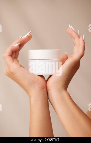 Mani eleganti con lunghe unghie bianche trattengono delicatamente un vaso bianco di crema cosmetica, che mostra un concetto di bellezza e cura della pelle contro un morbido dorso beige Foto Stock