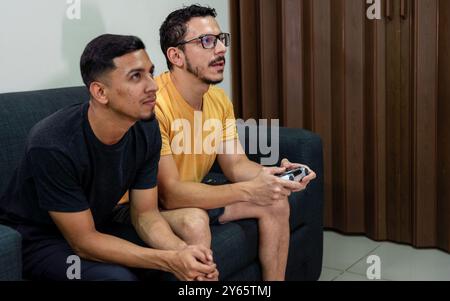 Una coppia gay, che fa parte della comunità LGBTQIA+, condivide un momento di divertimento mentre gioca ai videogiochi sulla loro console a casa, sottolineando un casual, in Foto Stock