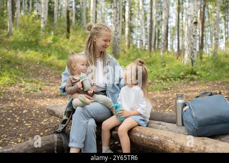 Una madre con i dreadlock e le sue due figlie si fanno una pausa su un tronco nel bosco, con una figlia che tiene un contenitore per bevande Foto Stock