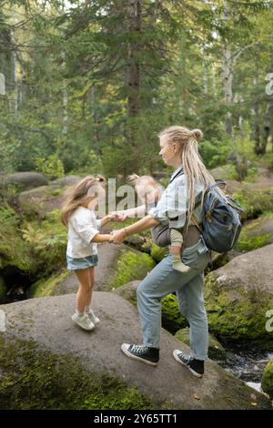 Una giovane madre con un portabicchieri e le sue due figlie sono viste camminare su rocce coperte di muschio in una foresta lussureggiante, simboleggiando un tempo di legame familiare Foto Stock