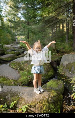 Una giovane ragazza si bilancia su una roccia ricoperta di muschio nella foresta con le braccia tese, esplorando la natura con la madre in una giornata di sole Foto Stock