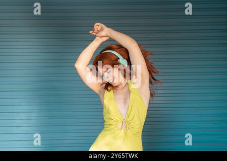 Una vivace donna dai capelli rossi con un abito giallo ama la musica attraverso le cuffie, ballando allegramente su uno sfondo blu testurizzato. Foto Stock