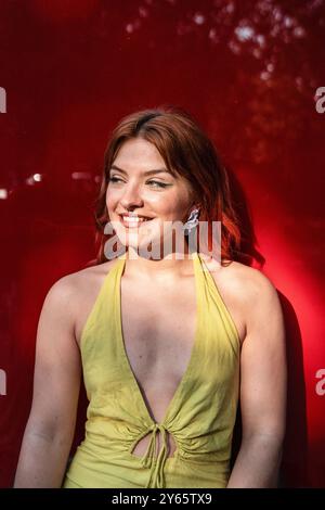 Una giovane donna con i capelli rossi sorride dolcemente alla luce del sole su un vivace sfondo rosso, indossando un abito giallo estivo con uno scollo profondo. Foto Stock