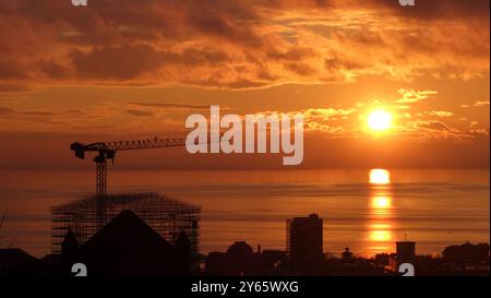 Una gru da costruzione torreggia sopra una struttura emergente, sagomata dal caldo bagliore di un sole che sorge su un mare calmo. Foto Stock