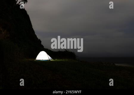 Una tenda solitaria si illumina sotto un cielo notturno, piazzata vicino al bordo di una scogliera panoramica, offrendo una serena fuga nella natura. L'immagine cattura l'essenza Foto Stock