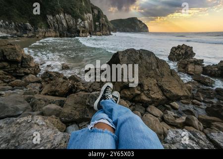 Le gambe rivestite di jeans e sneakers si estendono su una costa rocciosa frastagliata di fronte a un mare tranquillo sotto il cielo del tramonto, offrendo un momento sereno ed elegante di r Foto Stock