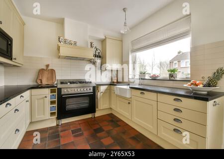 Una cucina ben illuminata con armadietti color crema, ripiani neri, piastrelle in terracotta ed elettrodomestici moderni. Foto Stock