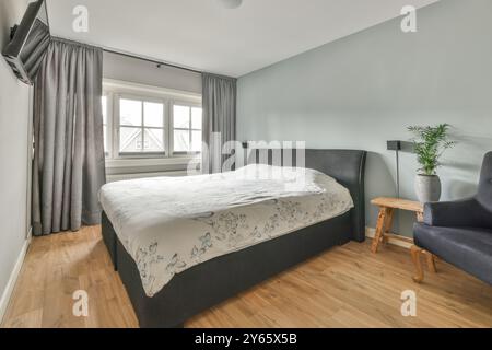 Una moderna camera da letto ben illuminata con un grande letto con una copertura a motivi floreali, pavimenti in legno duro, lunghe tende e una comoda poltrona. Foto Stock