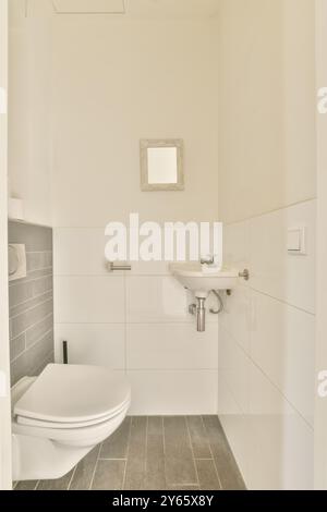 Questa immagine presenta un bagno moderno e minimalista con WC bianco, lavandino, pareti piastrellate e un piccolo specchio. Foto Stock