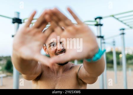 Un giovane uomo in forma sta realizzando una cornice con le mani intorno alla macchina fotografica in una palestra esterna di calisteni, mostrando un momento di divertimento durante una sessione di allenamento Foto Stock
