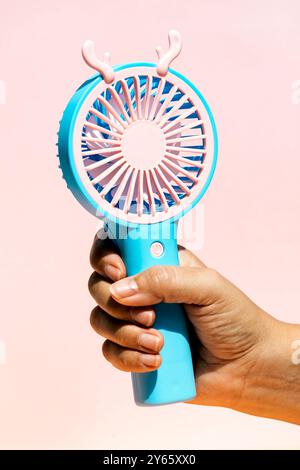 Immagine ravvicinata di una mano che afferra un bizzarro ventilatore portatile blu con stravagante corna rosa su uno sfondo rosa tenue, che illustra un divertimento e un divertimento Foto Stock