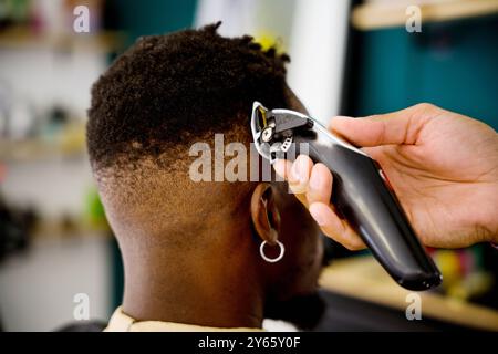 Immagine ravvicinata di un barbiere professionista, che utilizza i regolacapelli elettrici per modellare i capelli di un uomo nero in un barbiere a tema costiero Foto Stock