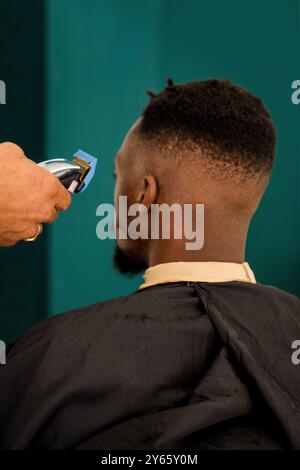 Una vista ravvicinata di un barbiere che taglia i capelli di un uomo nero in un ambiente barbiere verde vivace, mostrando abilità nello styling e nella cura dei capelli Foto Stock