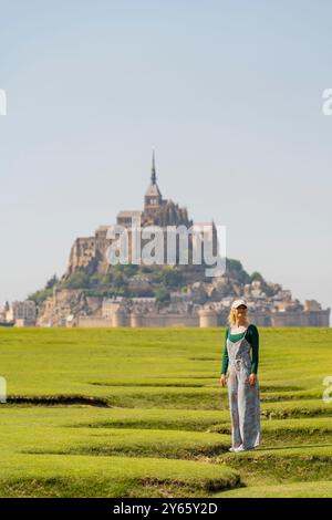 Una giovane donna vestita con una tuta si erge sulla palude verde con l'iconico Mont Saint-Michel sullo sfondo sotto un cielo azzurro Foto Stock