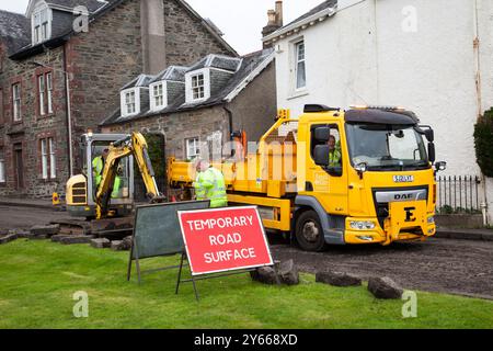 Lavoratori che riparano il fondo stradale, Rhu, Scozia, che mostrano camion e piccola scavatrice con cartello Temporary Road Surface (superficie stradale temporanea). Foto Stock