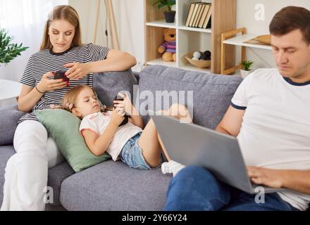 Famiglia a casa che utilizza laptop e smartphone seduti sul divano, divertiti online ossessionati Foto Stock