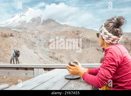 Una donna sola sedeva e beveva e guardava la coppia di turisti felici che camminavano in un bar panoramico sulle montagne. Panorama della cima del monte Kazbek nella nazionale di Kazbegi Foto Stock