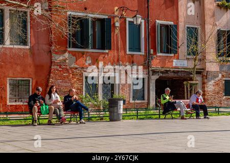 Le persone sedute sulle panchine godono del sole primaverile in campo San Giacomo, una piazza nel sestiere di Santa Croce a Venezia Foto Stock