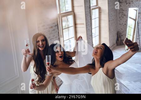 Le ragazze impazziscono. Vista dall'alto della giovane sposa e delle due damigelle che fanno i volti mentre scattano selfie in camerino Foto Stock