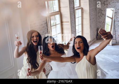 Mi sento così felice! Vista dall'alto della giovane sposa e delle due damigelle che fanno i volti mentre scattano selfie in camerino Foto Stock