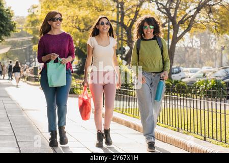 Un gruppo di tre donne che passeggiano felicemente all'aperto, trasportano borse della spesa in una giornata di sole, indossano abiti casual e occhiali da sole, e si godono il loro tim Foto Stock