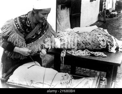 Una donna Magyar slega il suo bambino in modo che possa arrotolarlo nel cuscino in cui è stato trasportato. È tradizione portare bambini legati nelle culle in Ungheria. Mesokovesd, Ungheria - 1925 Foto Stock