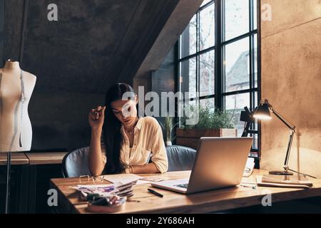 Trasformare le idee in abbigliamento. Bella giovane donna che lavora su schizzi mentre è seduta nel suo laboratorio Foto Stock