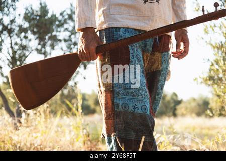 Uno scatto ravvicinato di un uomo di 40 anni che tiene in mano un baglama mentre cammina attraverso un campo d'ulivo, indossando pantaloni colorati a motivi geometrici e una camicia leggera Foto Stock