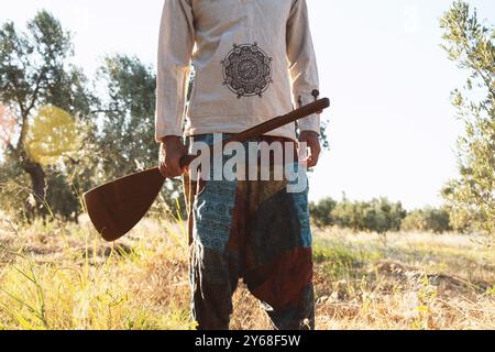 Uno scatto ravvicinato di un uomo di 40 anni che tiene in mano un baglama mentre cammina attraverso un campo d'ulivo, indossando pantaloni colorati a motivi geometrici e una camicia leggera Foto Stock