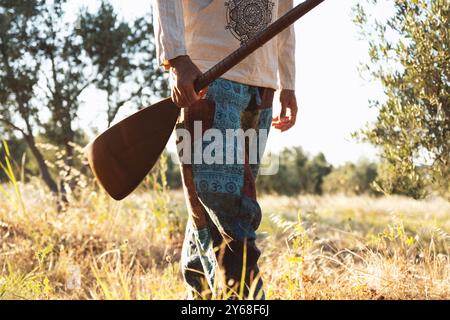 Uno scatto ravvicinato di un uomo di 40 anni che tiene in mano un baglama mentre cammina attraverso un campo d'ulivo, indossando pantaloni colorati a motivi geometrici e una camicia leggera Foto Stock