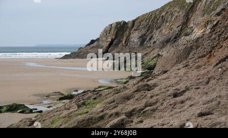 Un tranquillo paesaggio costiero caratterizzato da aspre scogliere e una spiaggia sabbiosa con dolci onde sotto un cielo nuvoloso. Foto Stock