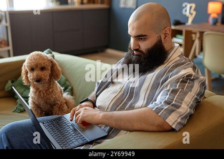 L'uomo barbuto seduto sul divano scrivendo su un computer portatile mentre un piccolo cane guarda attentamente accanto, creando un'atmosfera accogliente e concentrata in un moderno soggiorno Foto Stock