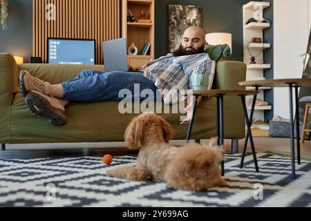 Uomo sdraiato sul divano con un computer portatile in grembo, che lavora a distanza in un accogliente soggiorno con mobili moderni, scaffale e adorabile cane Foto Stock