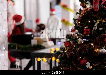 Primo piano sull'albero di Natale decorato con stile e i dipendenti ricevono regali speciali da Babbo Natale durante le vacanze invernali su sfondo sfocato. Il manager in costume di Natale regala regali ai lavoratori Foto Stock