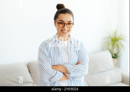 Ritratto di una donna indiana o araba sicura di sé, intelligente e bella con gli occhiali, manager di successo, seo digitale, web designer, stare in casa con le braccia incrociate, guardare la fotocamera, sorridere Foto Stock