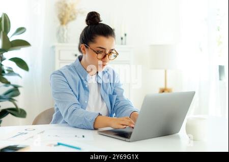 Donna intelligente e concentrata di nazionalità araba o indiana con occhiali, impiegato, manager digitale, guarda lo schermo del notebook, cerca informazioni per un progetto, digita la tastiera Foto Stock