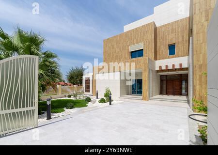 Moderna entrata in villa ad Abu Dhabi, caratterizzata da una facciata in pietra chiara e legno, elegante porta bianca, vialetto pavimentato e lussureggiante giardino con palme Foto Stock