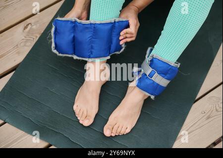 Primo piano di una donna che regola i pesi alla caviglia blu su un tappetino da yoga, pronto per un allenamento fitness all'aperto. Ideale per l'allenamento di forza, l'esercizio fisico e la guarigione Foto Stock