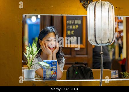 Ragazza con la testa tra le mani, sorridente e guardando fuori dalla finestra di notte - Hoi An Foto Stock