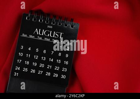Agosto 2025 calendario desktop a spirale nero su sfondo tessile rosso. Foto Stock