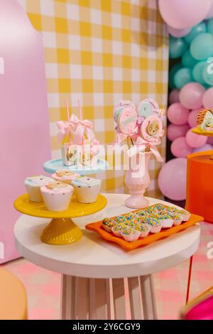 Tavolino da dessert colorato con cupcake, biscotti e tortini per una festa per bambini, con palloncini pastello e sfondo a scacchi. Foto Stock