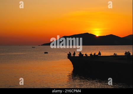 Sagome di pescatori con canne da pesca che pescano sul molo in mare la mattina all'alba in estate Foto Stock