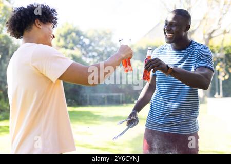 Brindisi con un drink, due diversi amici maschi si godono il barbecue all'aperto con le pinze in mano Foto Stock