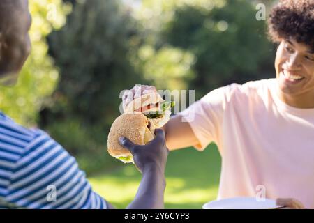Due diversi amici maschi si divertono all'aperto, condividono hamburger e sorridono in giardino Foto Stock