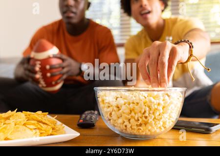 Primo piano, a casa, a guardare il calcio, due diversi amici maschi che mangiano popcorn e patatine fritte e si divertono Foto Stock