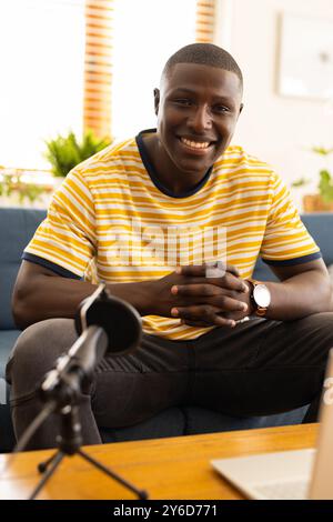 A casa, sorridente uomo afroamericano seduto sul divano con microfono e laptop, godendo conversa Foto Stock